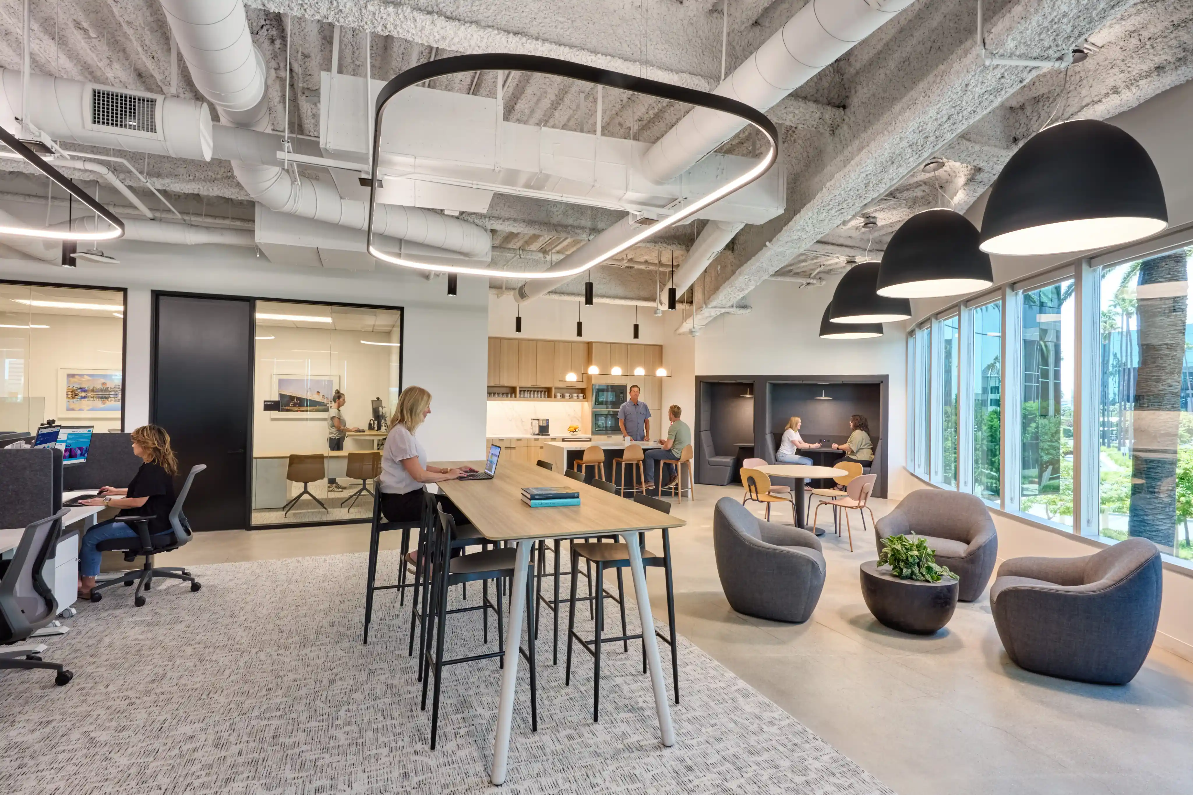 Modern open office space with natural light, featuring people working at desks and high tables. Cozy seating area has round chairs and greenery.