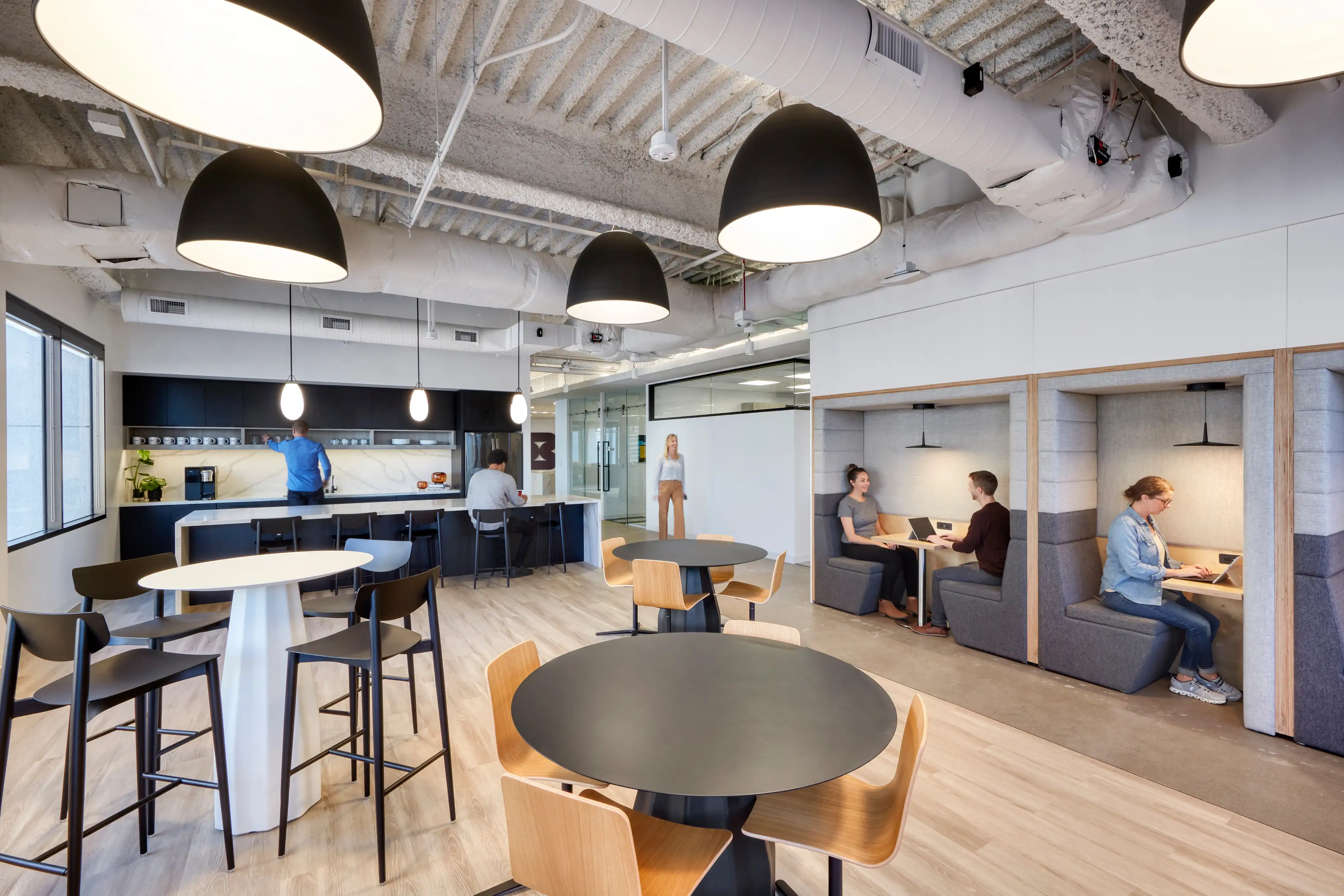 Modern office kitchen and lounge with black pendant lights, round tables, and cozy seating nooks. People work and socialize, creating a collaborative atmosphere.