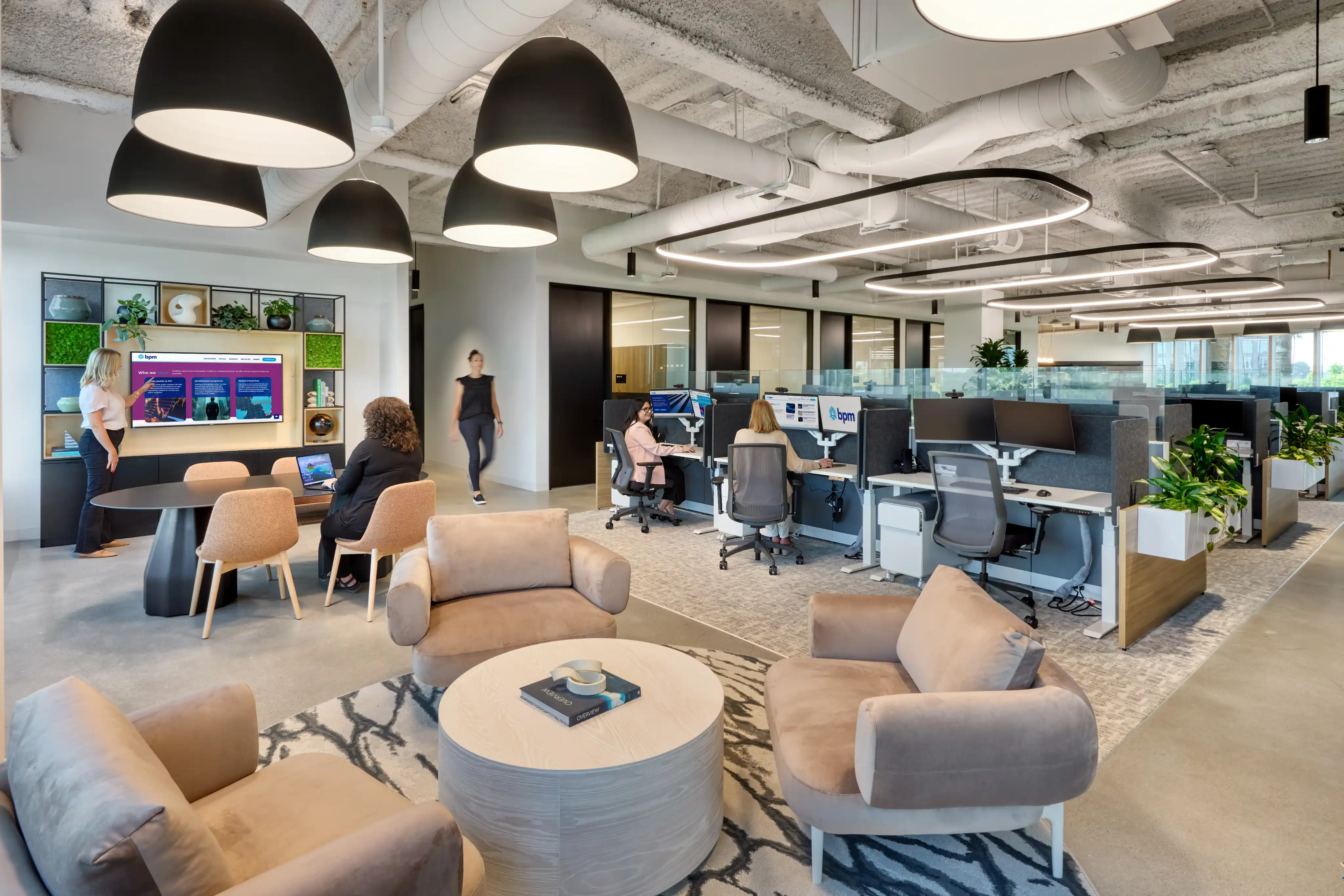 Modern office space with an open layout, featuring cozy seating, a round table with laptops, plants, large lamps, and employees working at desks.