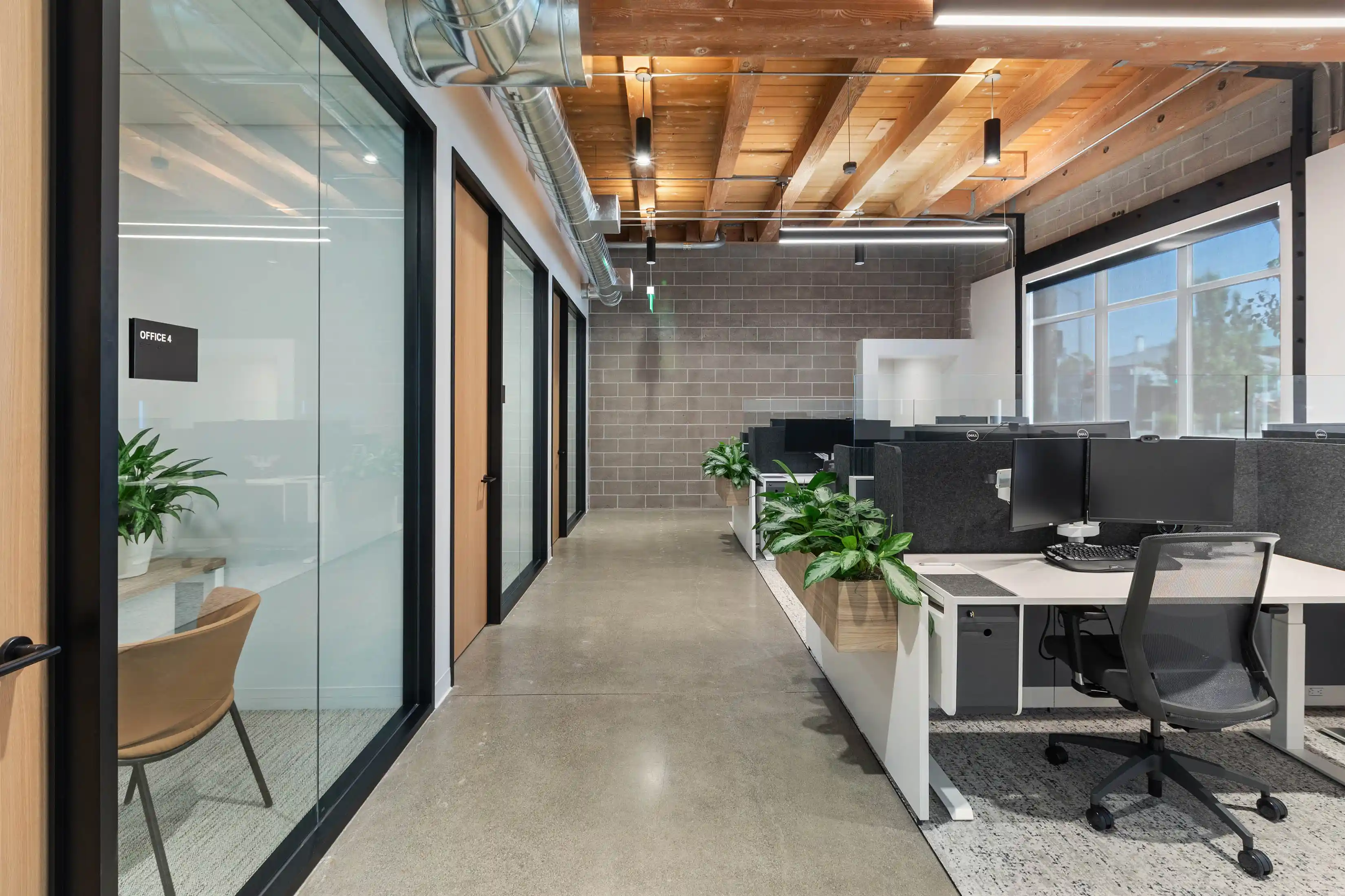 Modern office interior with concrete floors, glass-walled private offices on the left, and open cubicles with computers on the right. Natural light and plants create a fresh, professional atmosphere.