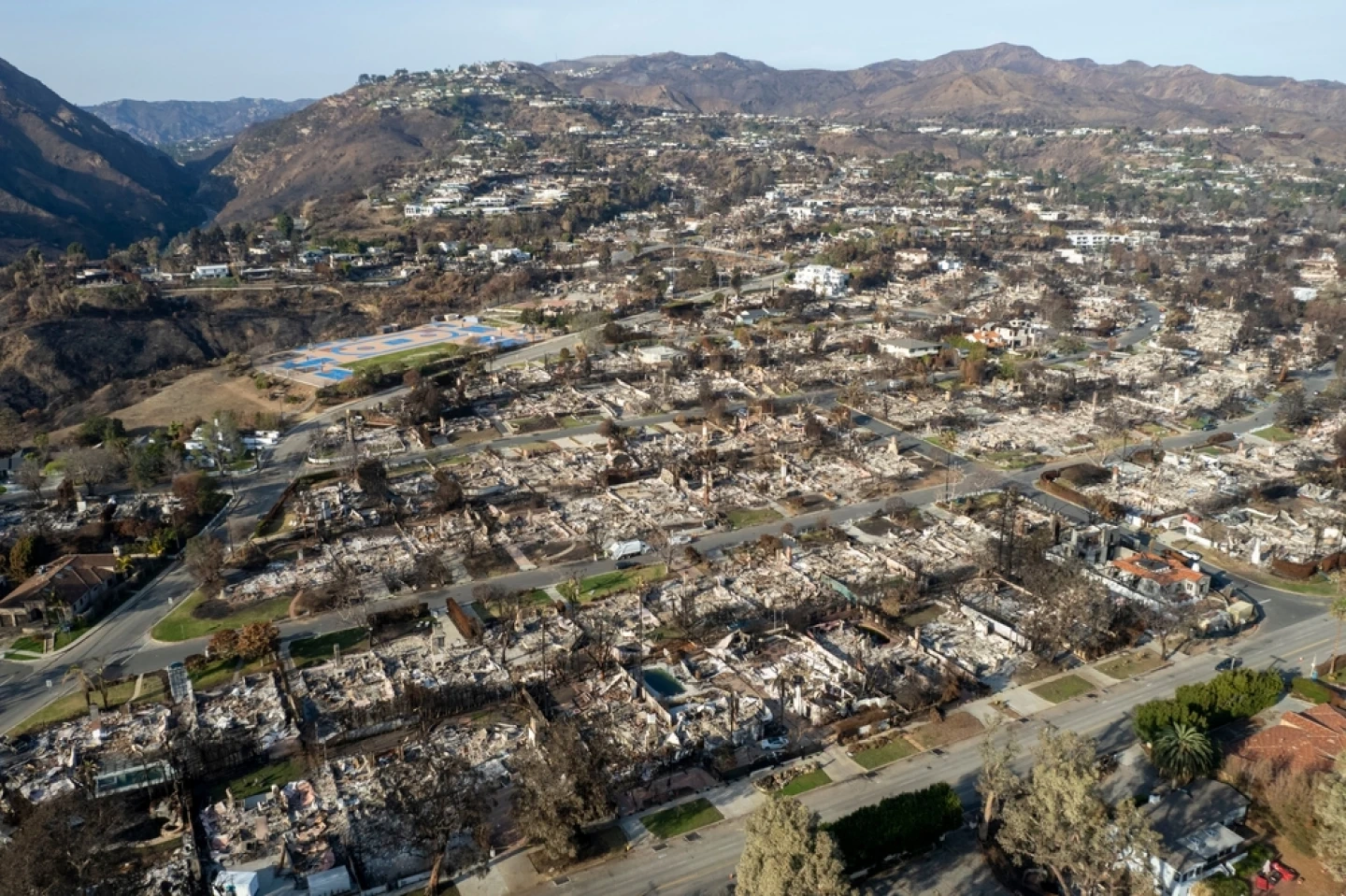 Aerial view of a neighborhood devastated by fire, showing rows of charred, collapsed homes amidst barren, brown hills. A somber, desolate scene.