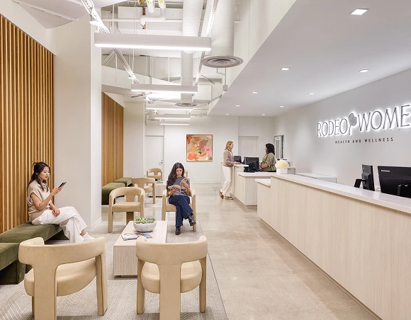 Modern clinic reception area with neutral tones. Three women seated, two using phones, one reading. Reception desk on right, two staff members assisting. Calming ambiance.