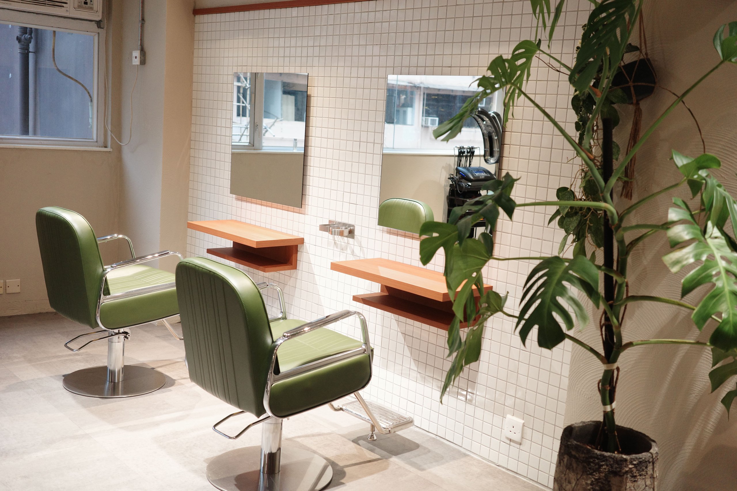 Modern salon interior with two green styling chairs, wall-mounted mirrors, wooden shelves, and a large potted plant.
