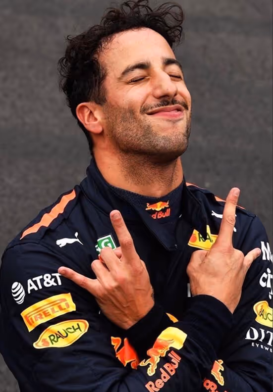 Man in a Red Bull racing suit smiling with eyes closed and making a hand gesture crossing his arms.