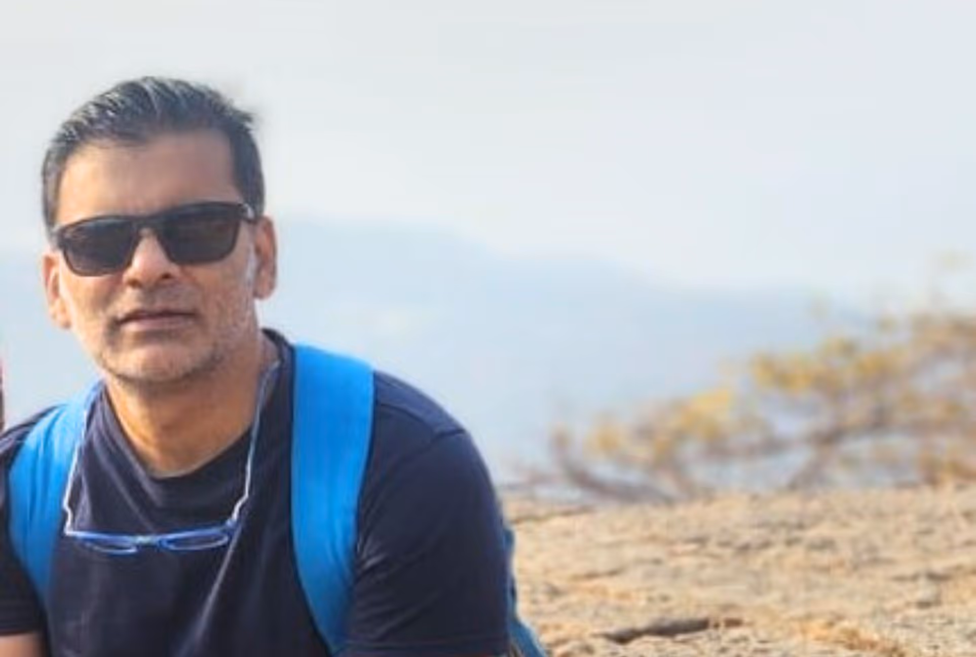 Man wearing sunglasses, a blue backpack, and a black shirt outdoors with a blurred natural background.