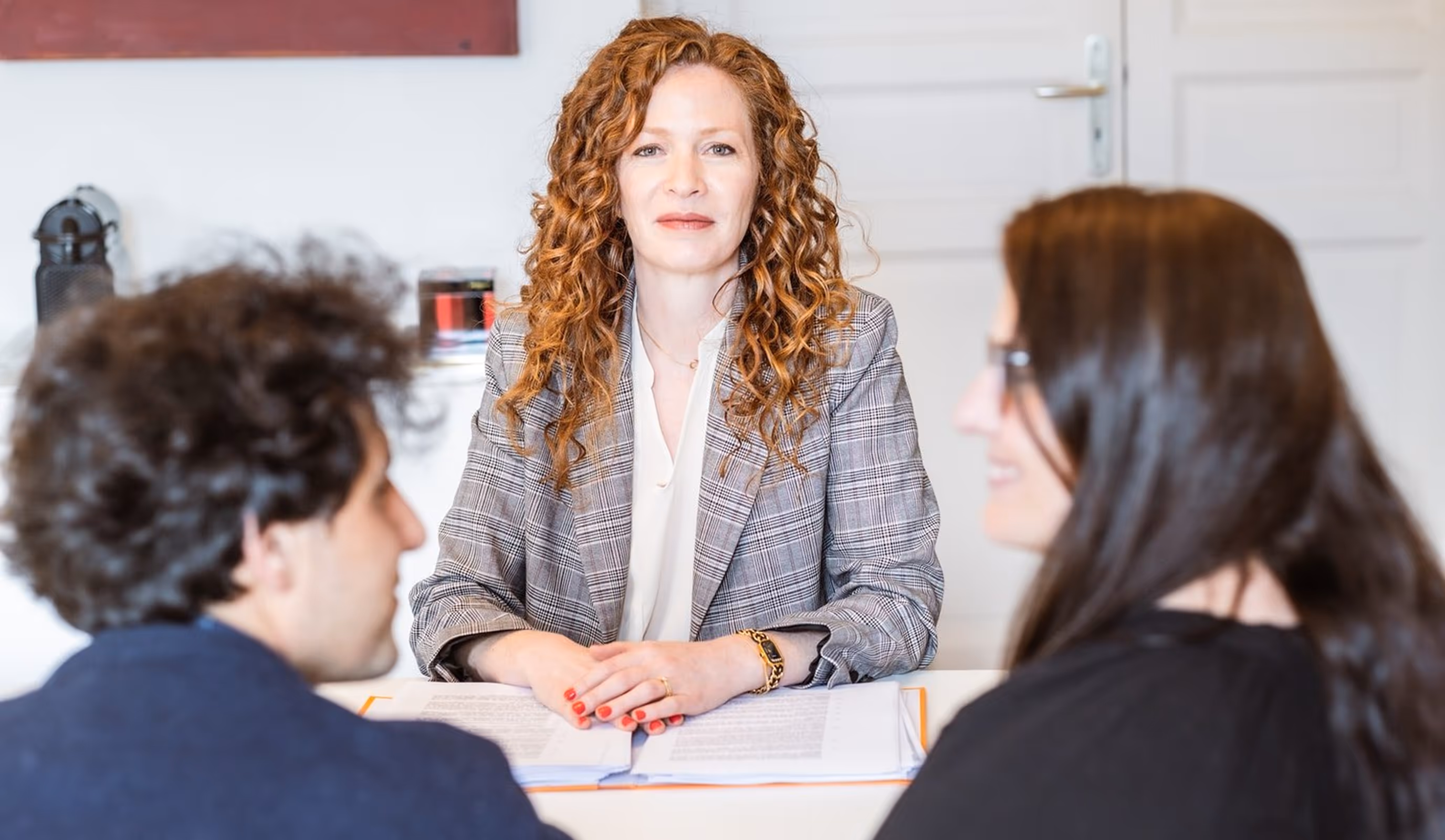 A woman with curly hair and a plaid blazer is sitting at a table with documents, facing two people in conversation.