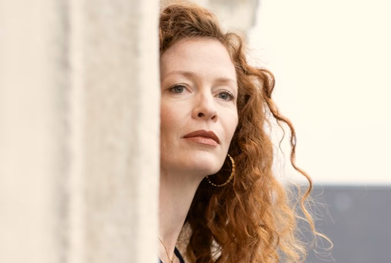Close-up of a woman with curly red hair and hoop earrings looking thoughtfully past a beige wall.