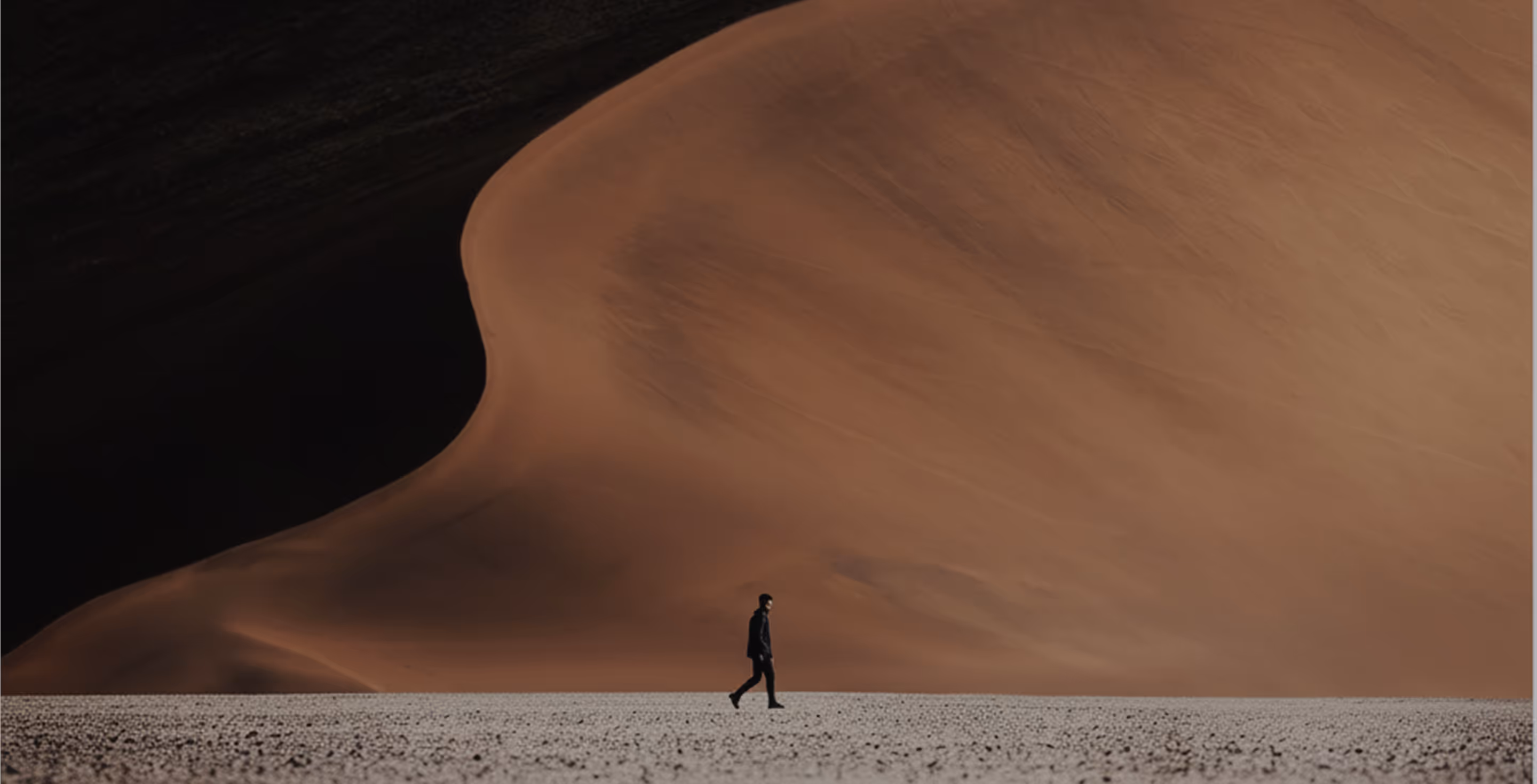 Person walking alone on a flat, barren desert plain with a massive sand dune in the background.