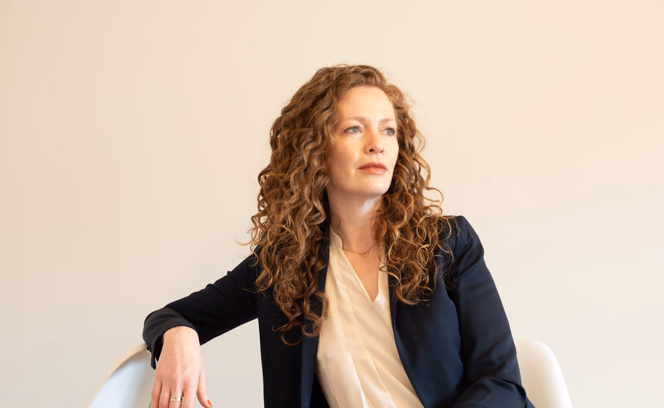 Woman with curly red hair wearing a black blazer and light blouse, sitting on a white chair looking thoughtfully to the side.