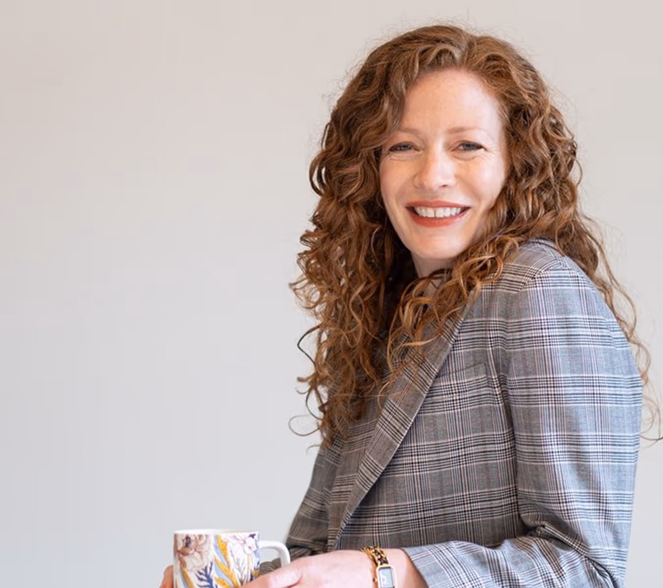 Smiling woman with curly red hair wearing a plaid blazer and holding a floral mug against a plain background.