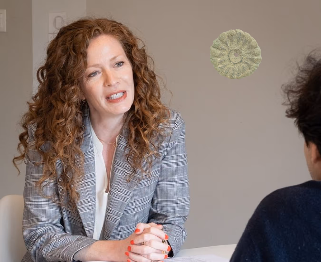 Woman with curly hair wearing a plaid blazer talking to a person across a table in a meeting.