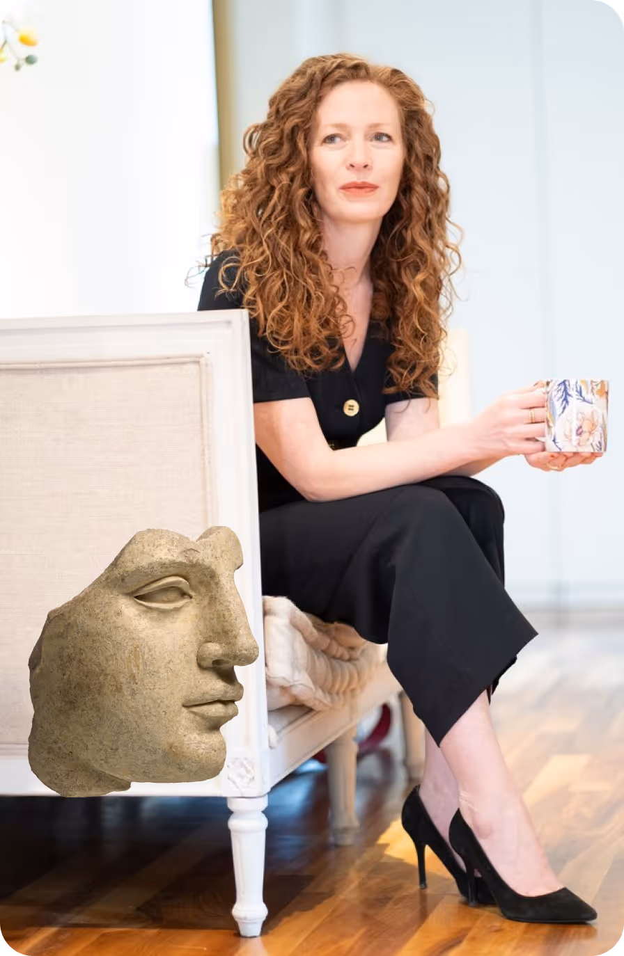 Red-haired woman in black outfit sitting on a white chair holding a floral mug, with a large stone sculpture of a face next to the chair.