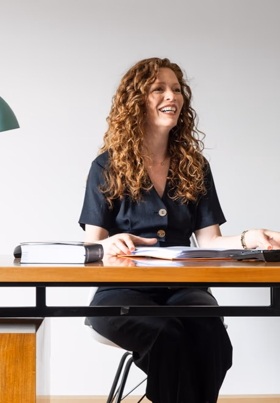Smiling woman with curly hair sitting at a desk with an open book and a closed thick book.
