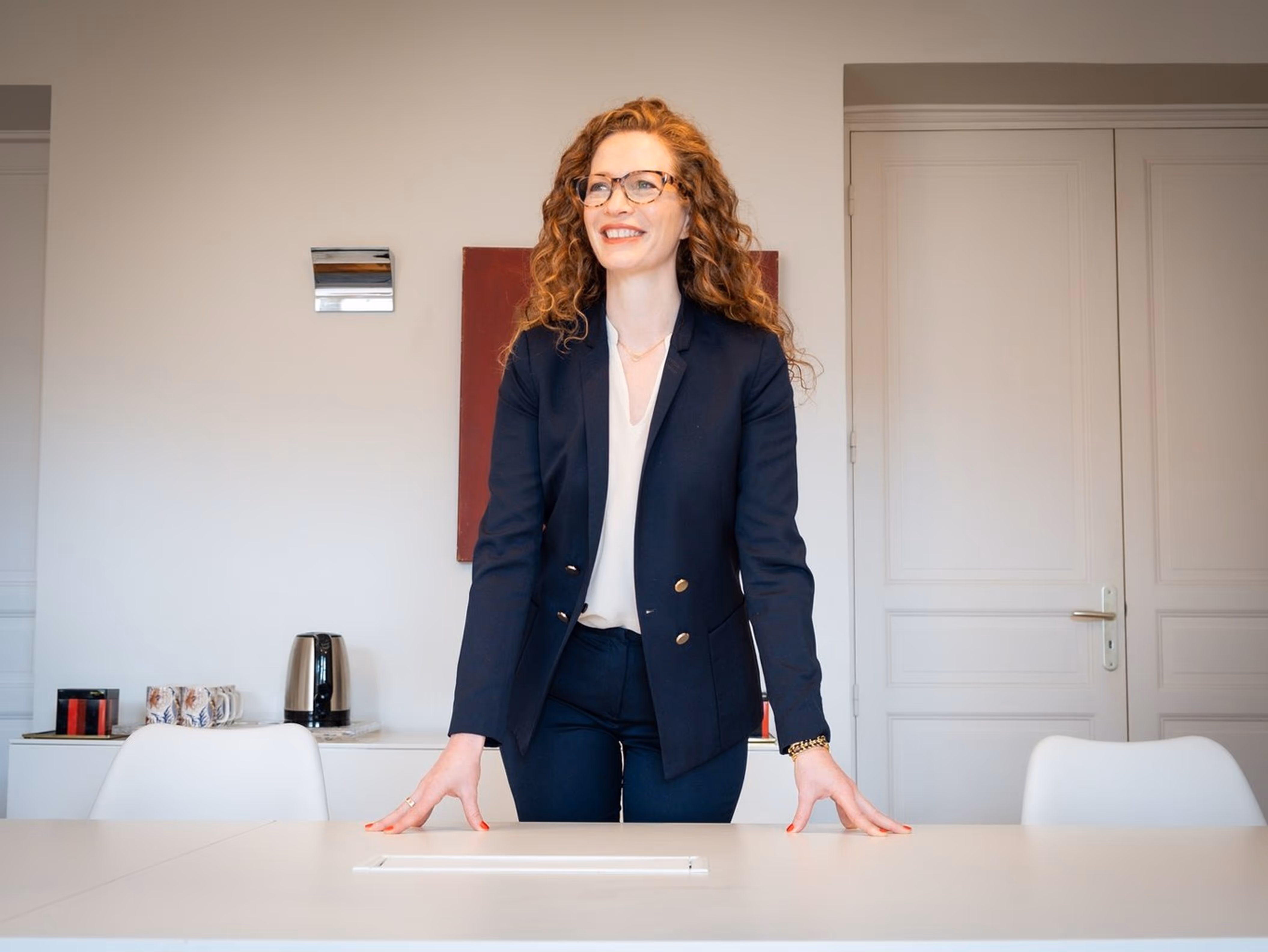 Smiling businesswoman with curly red hair and glasses standing behind a white desk in a modern office.