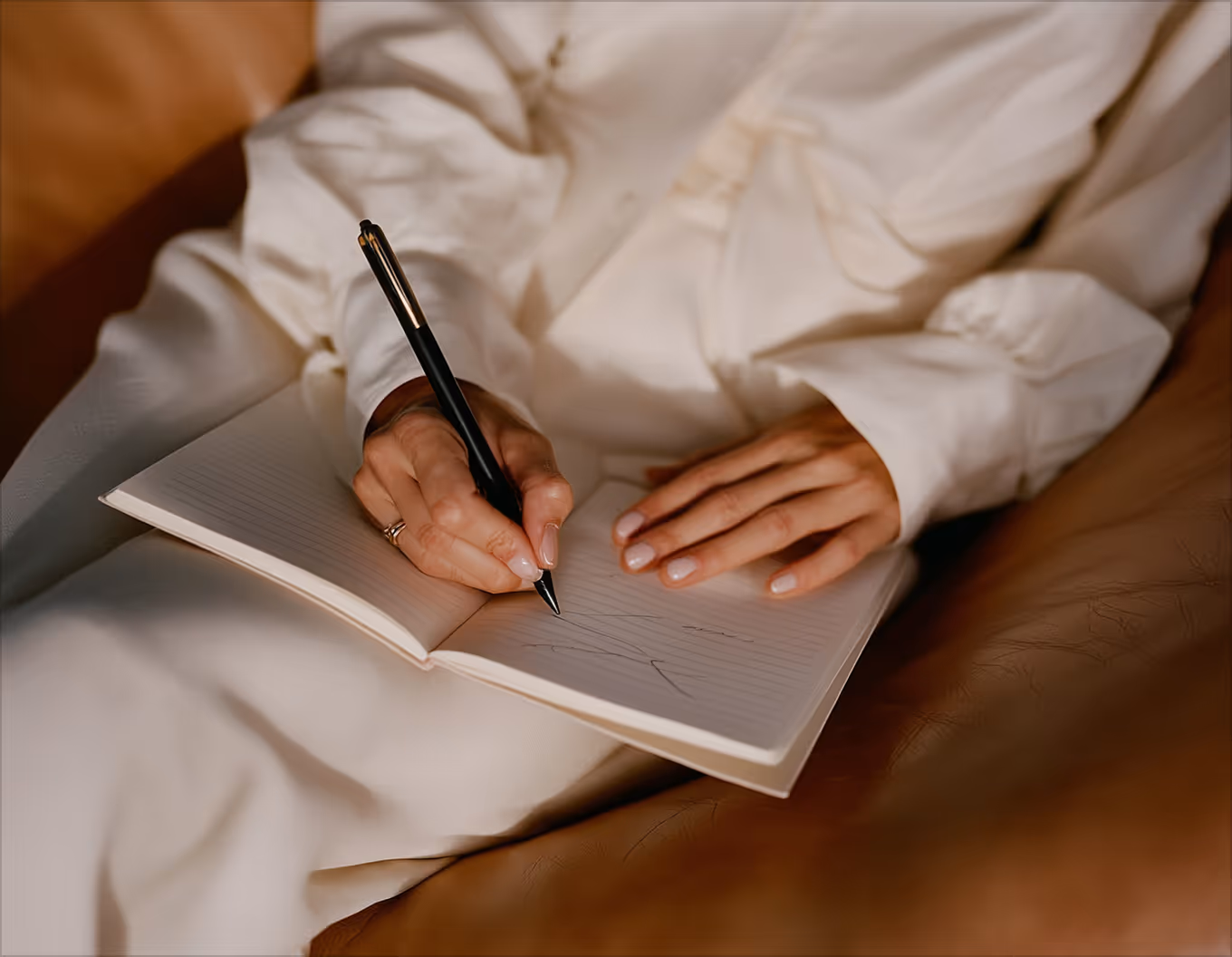 Person in white clothing writing in an open lined notebook with a black pen while seated on a brown leather couch.