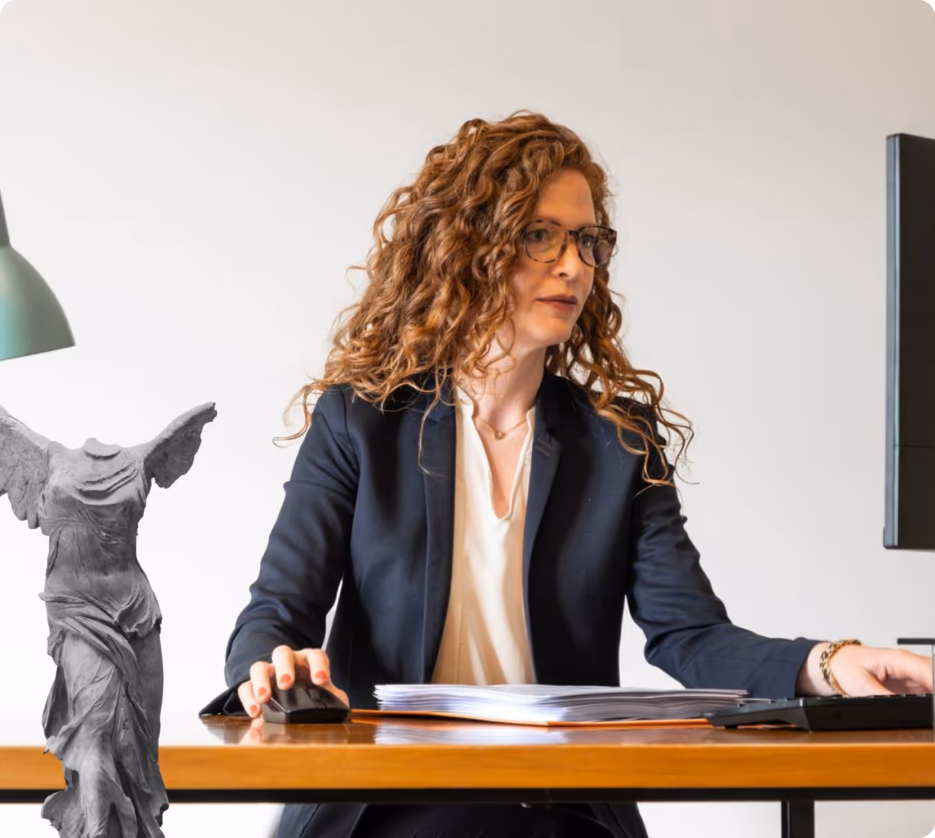 Woman with curly red hair and glasses working at a desk using a computer mouse and keyboard with a stack of papers in front of her and a small statue of a winged figure on the left.
