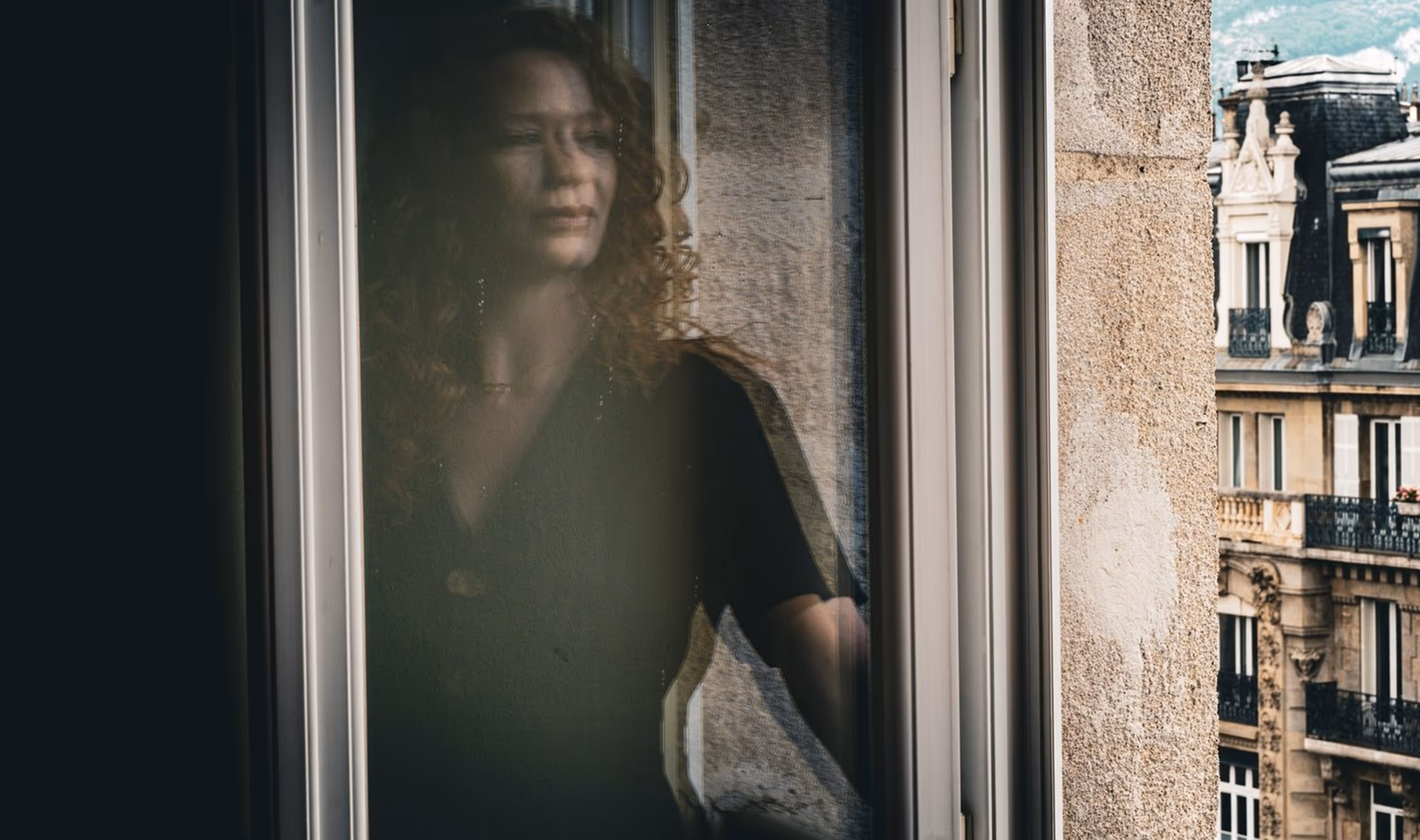 Woman with curly hair looking out a window with a reflection on the glass, historic buildings visible outside.