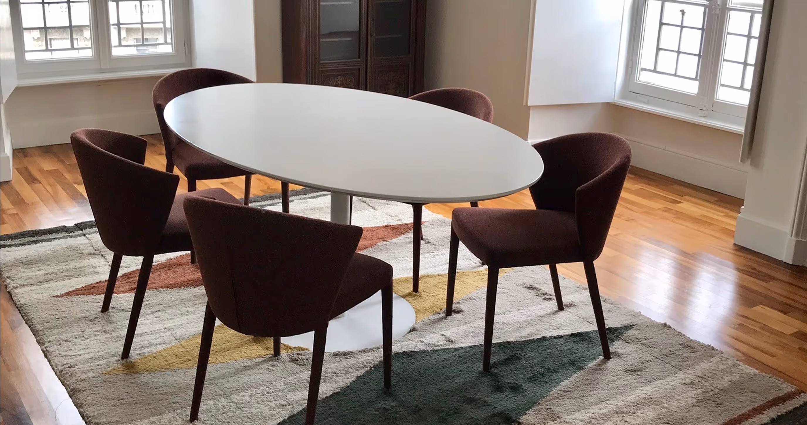 Modern oval white table surrounded by five dark brown upholstered chairs on a geometric patterned rug in a room with wooden floors and large windows.