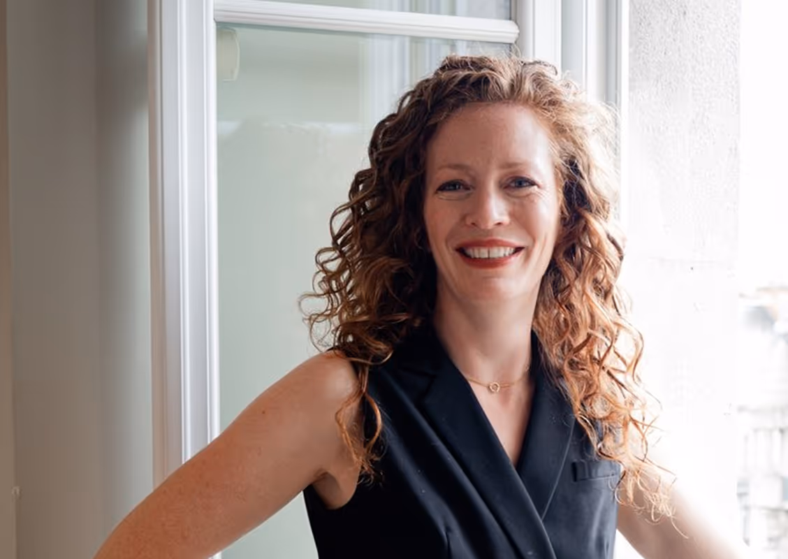 Smiling woman with curly red hair wearing a sleeveless black top standing by a window.