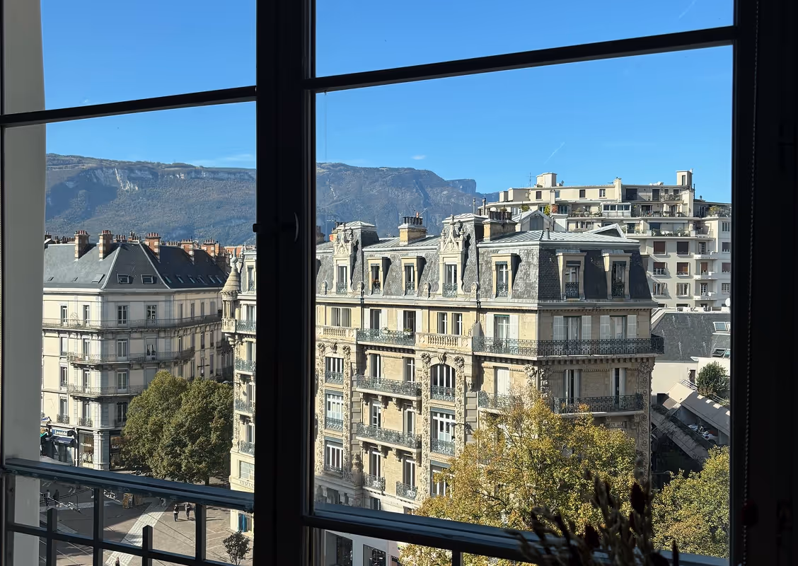 View through a window showing classic European buildings with mansard roofs, trees, and mountains in the background under a clear blue sky.