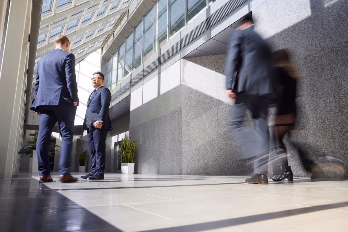 Two businessmen in suits standing and talking in a bright modern office hallway with large windows, while two blurred people walk past.