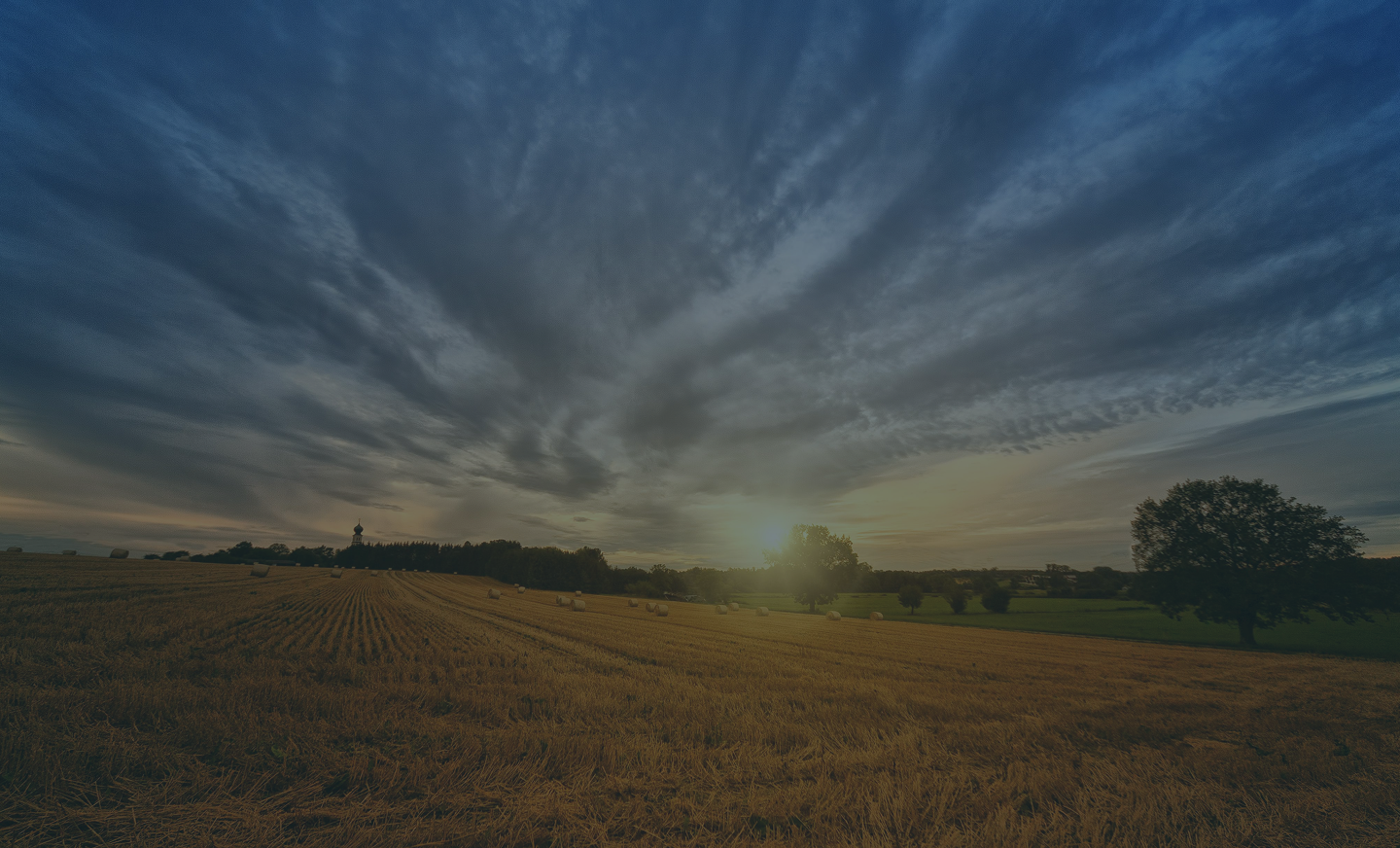 Sunset over a harvested field with hay bales, scattered trees, and a cloudy sky.