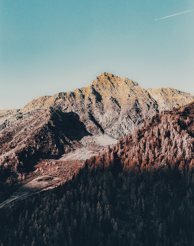 Rocky mountain peak with forested slopes under a clear blue sky with a small plane and contrail.