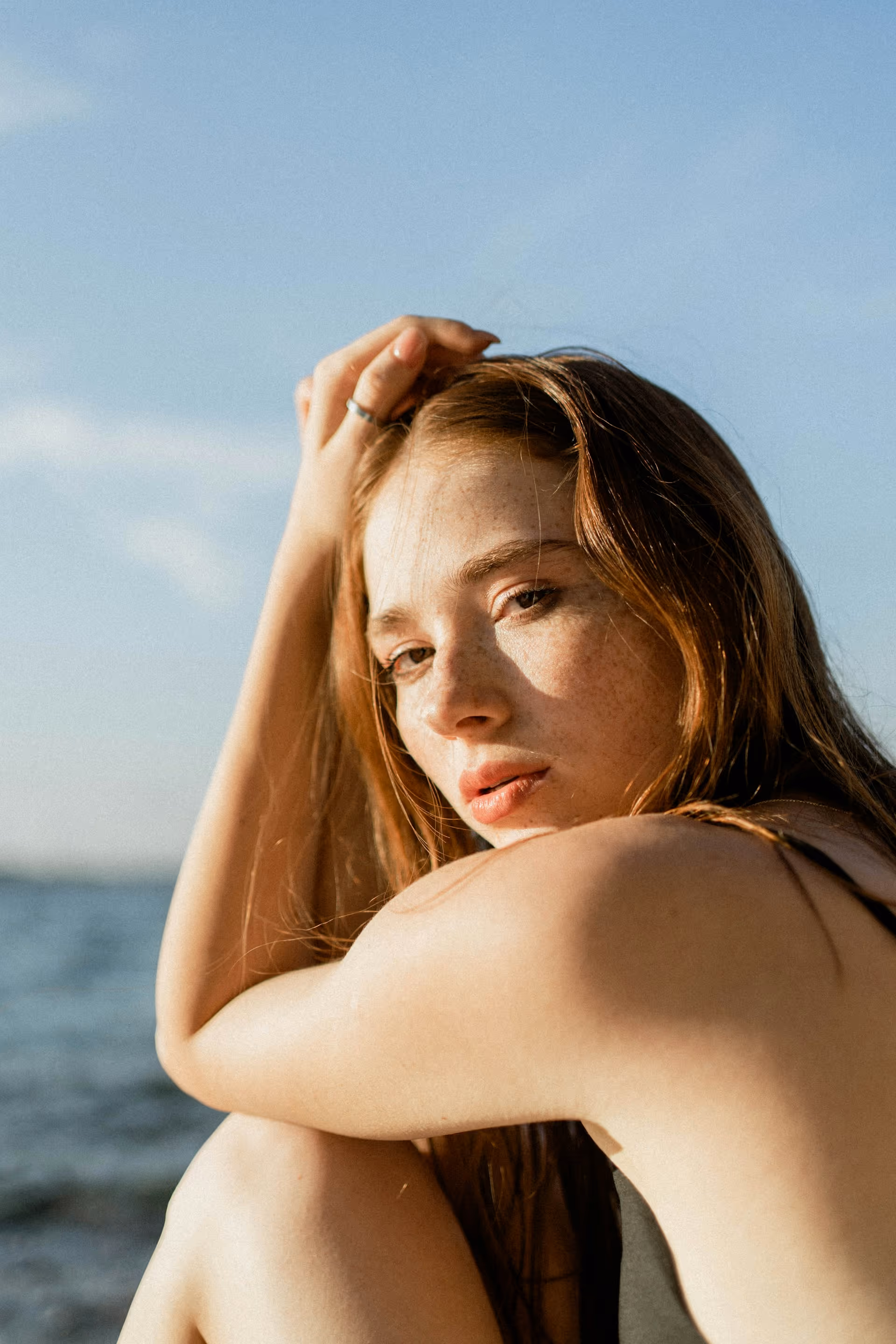 Close-up of a young woman with red hair and freckles sitting outdoors near water, resting her arm on her knee and looking softly at the camera.
