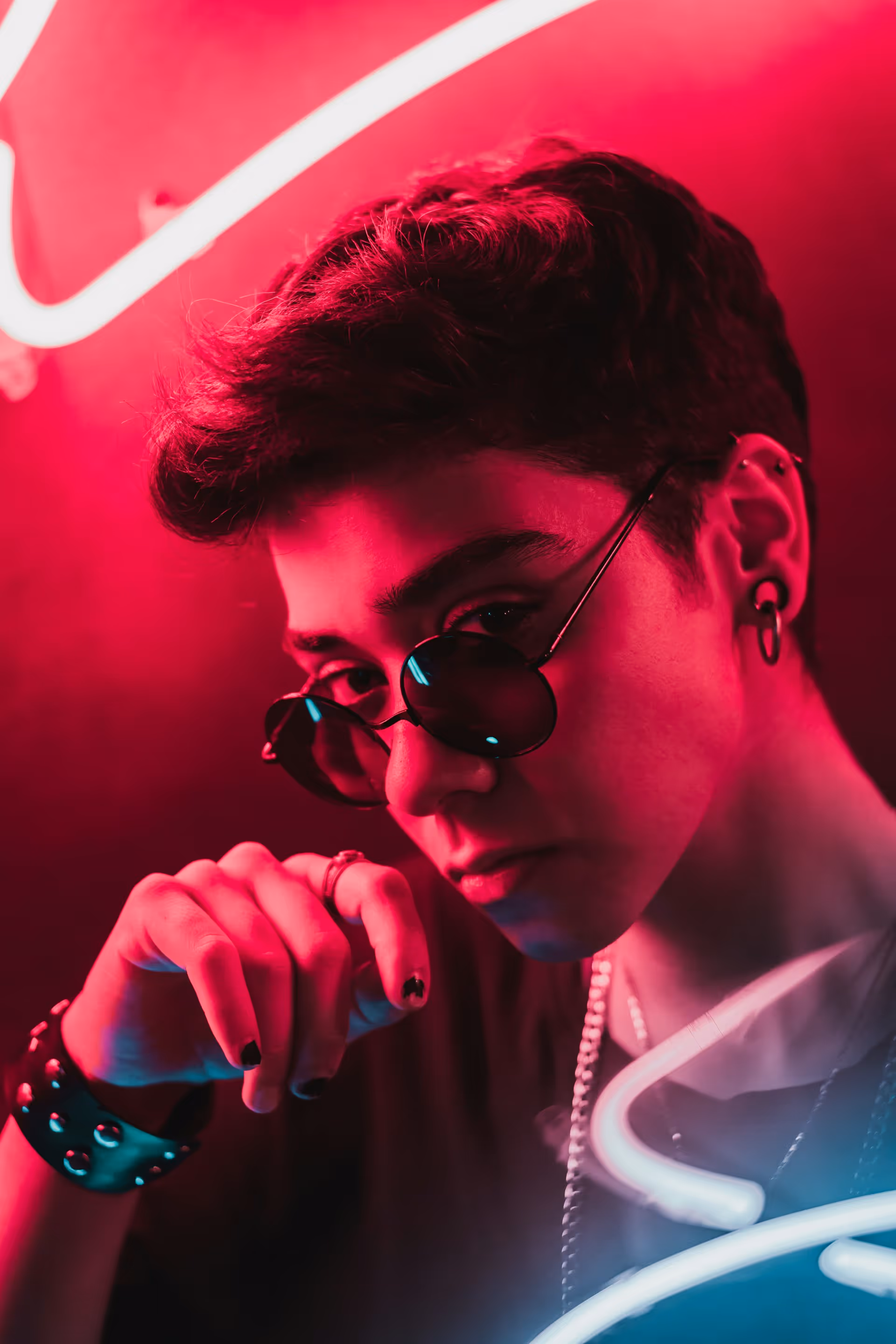 Young person with short hair wearing round sunglasses, black nail polish, and ear piercings, illuminated by pink and blue neon lights.