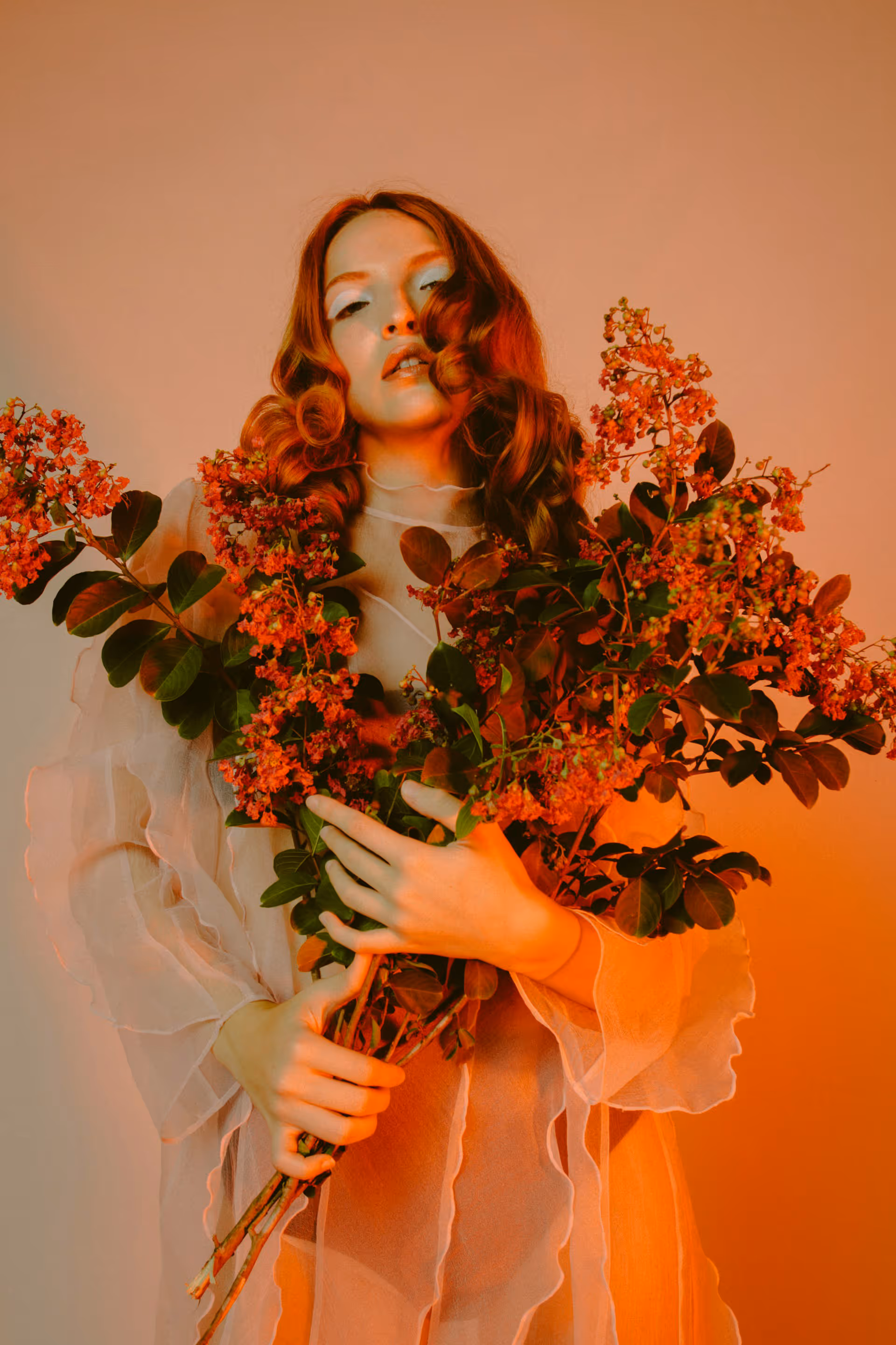 Woman with red curly hair wearing a sheer, ruffled dress holding a large bouquet of orange flowers and green leaves.
