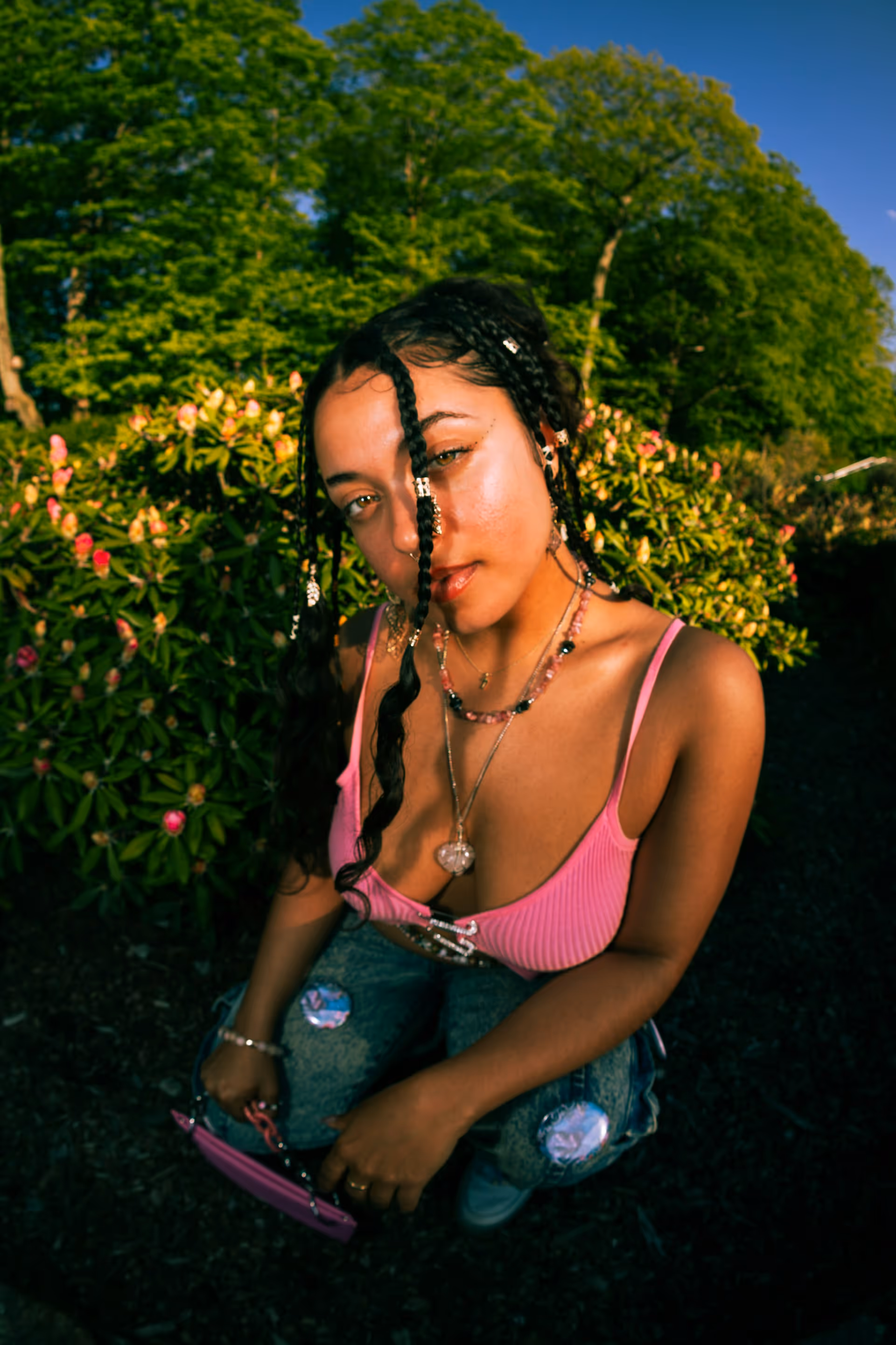 Young woman with braided hair adorned with beads, wearing a pink tank top and necklaces, crouching outdoors in front of green bushes with pink flowers.