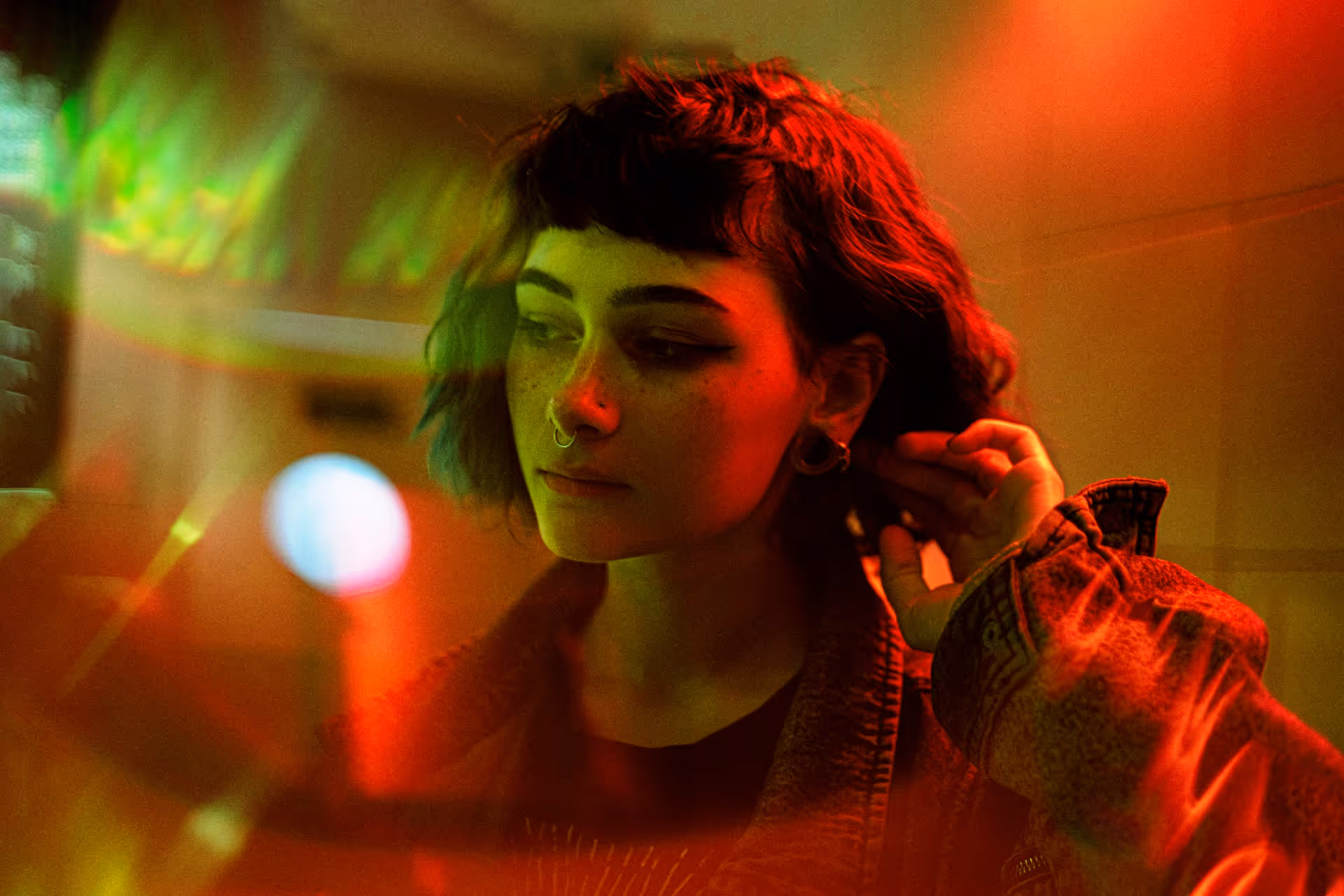 Young person with short dark hair, septum piercing, and multiple earrings looking down with red and green lighting effects.
