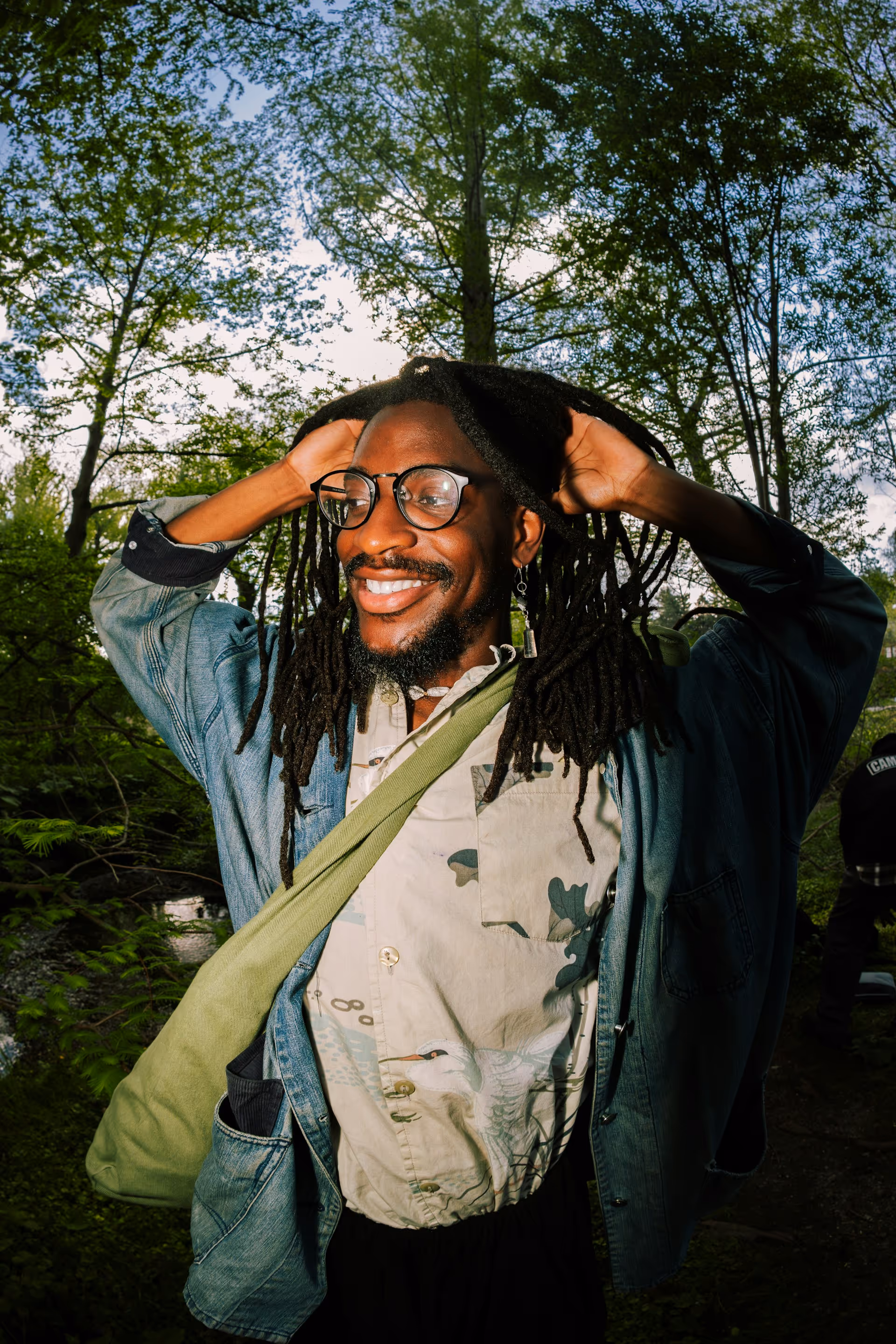 Smiling man with dreadlocks and glasses standing outdoors among green trees with hands behind his head.