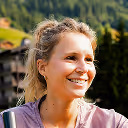 Smiling woman with blonde hair outdoors with blurred green trees and wooden buildings in the background.