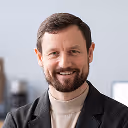 Smiling man with short brown hair and beard wearing a beige turtleneck and dark blazer against a light background.