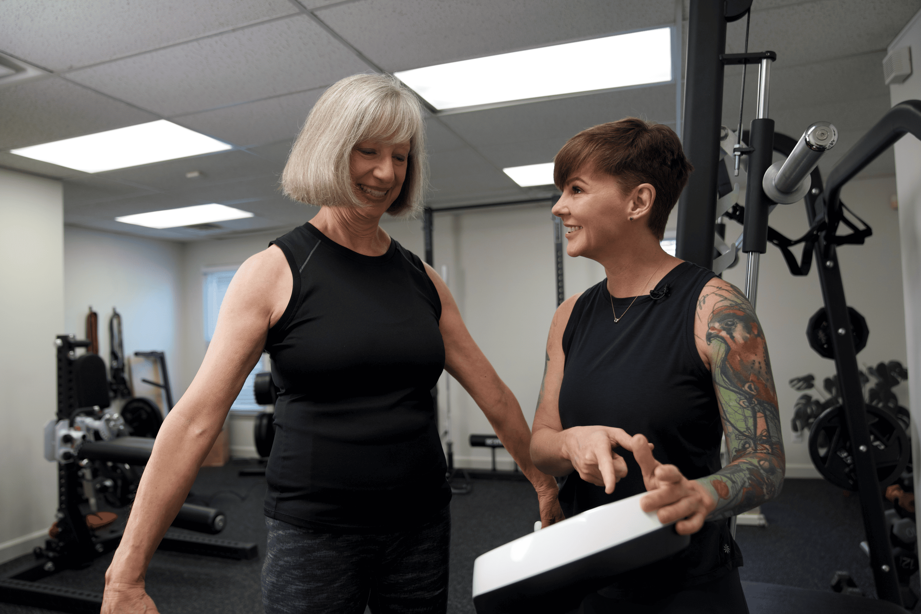 Female fitness trainer with a tattooed arm talking to and assisting an older woman in a gym.