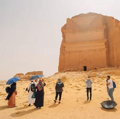 مجموعة من السياح يحملون مظلات ويزورون موقعًا أثريًا صخريًا في الصحراء تحت سماء صافية.