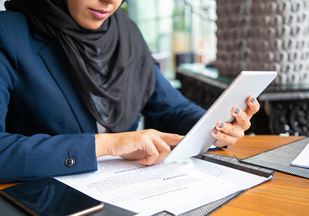 امرأة ترتدي الحجاب والبدلة الزرقاء تستخدم جهاز لوحي وهي تجلس على مكتب وترتبط بوثيقة.