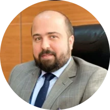 رجل يرتدي بدلة رمادية مع ربطة عنق زرقاء جالسا على كرسي مكتبي أسود أمام خلفية خشبية.