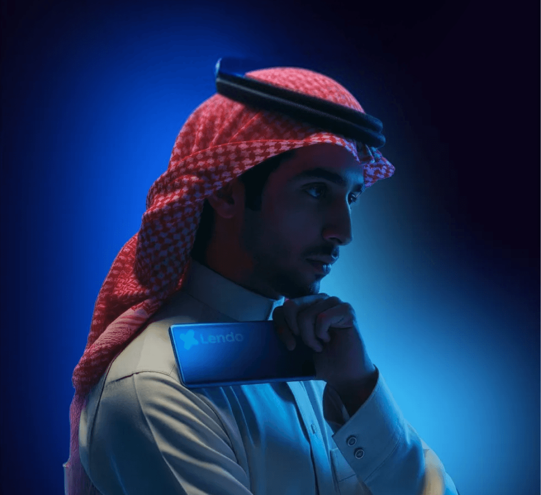 رجل يرتدي ثوباً عربيًا وغترة حمراء البيضاء يحمل هاتفًا يظهر شعار Lendo بخلفية إضاءة زرقاء.
