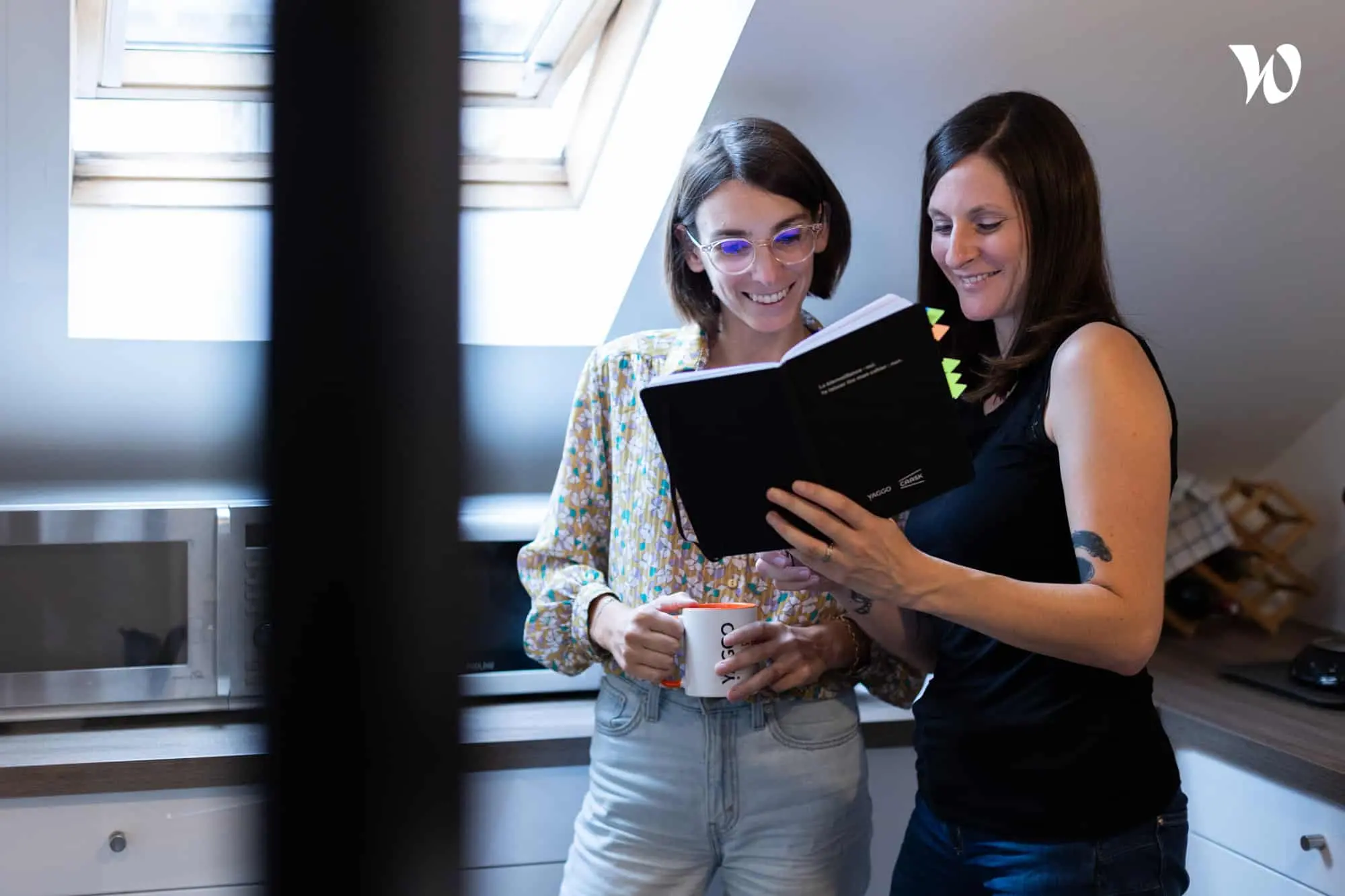 Deux femmes souriantes lisant un cahier noir ensemble dans une cuisine contemporaine.