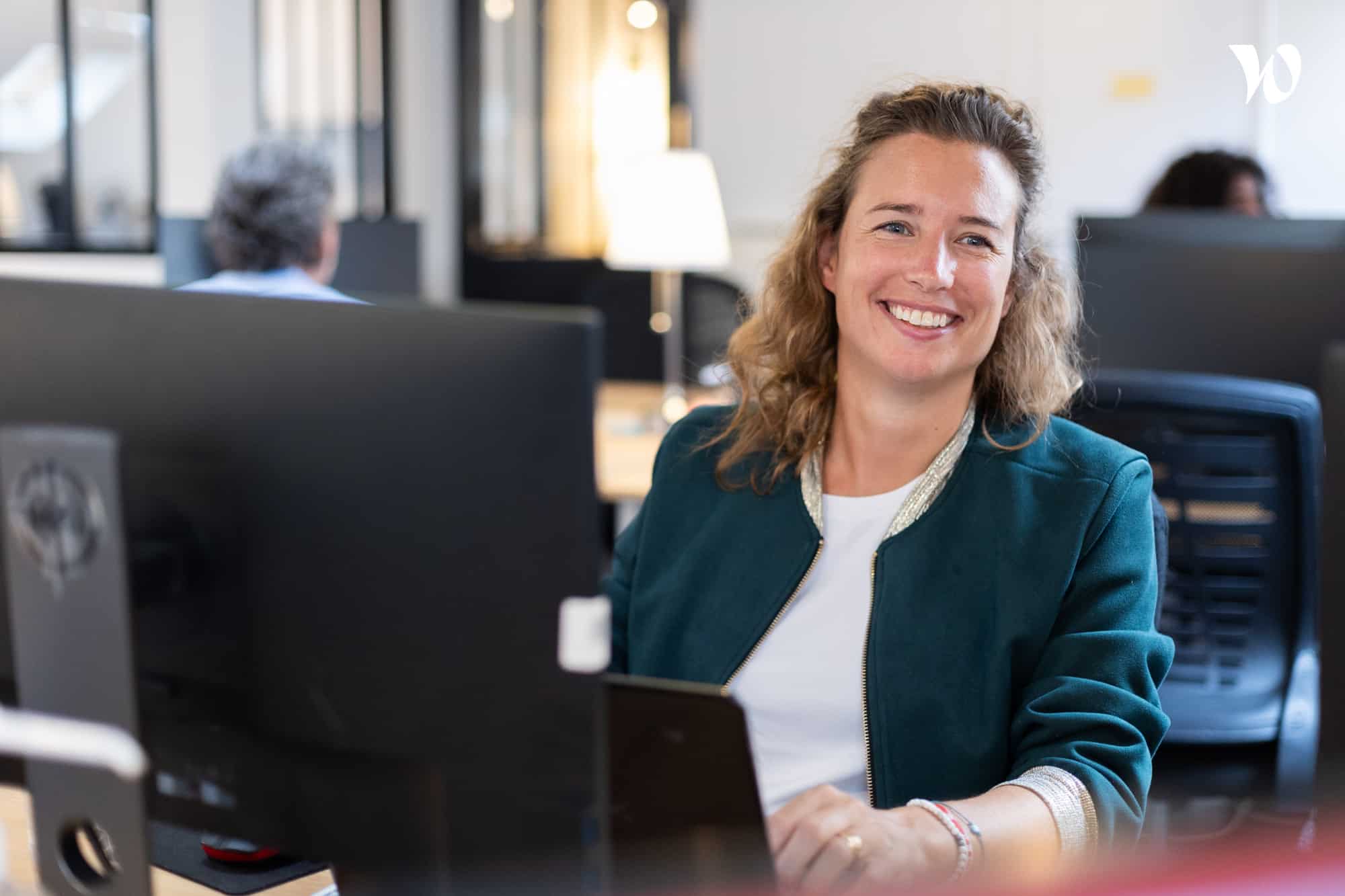 Femme souriante assise à un bureau devant un ordinateur dans un bureau moderne.