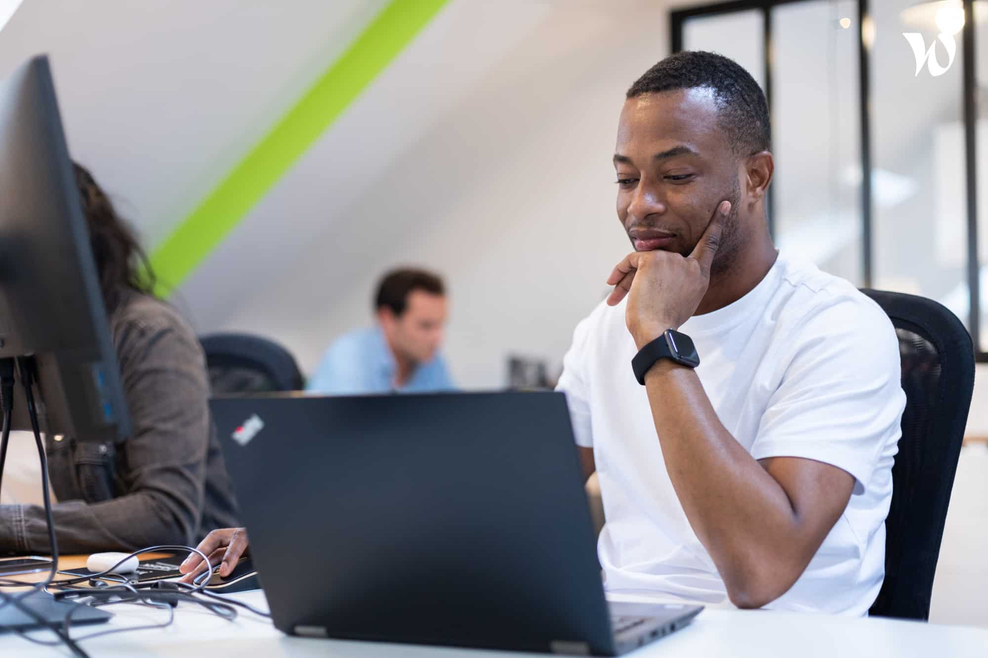 Un homme souriant regarde un ordinateur portable dans un bureau moderne avec d'autres collègues en arrière-plan.