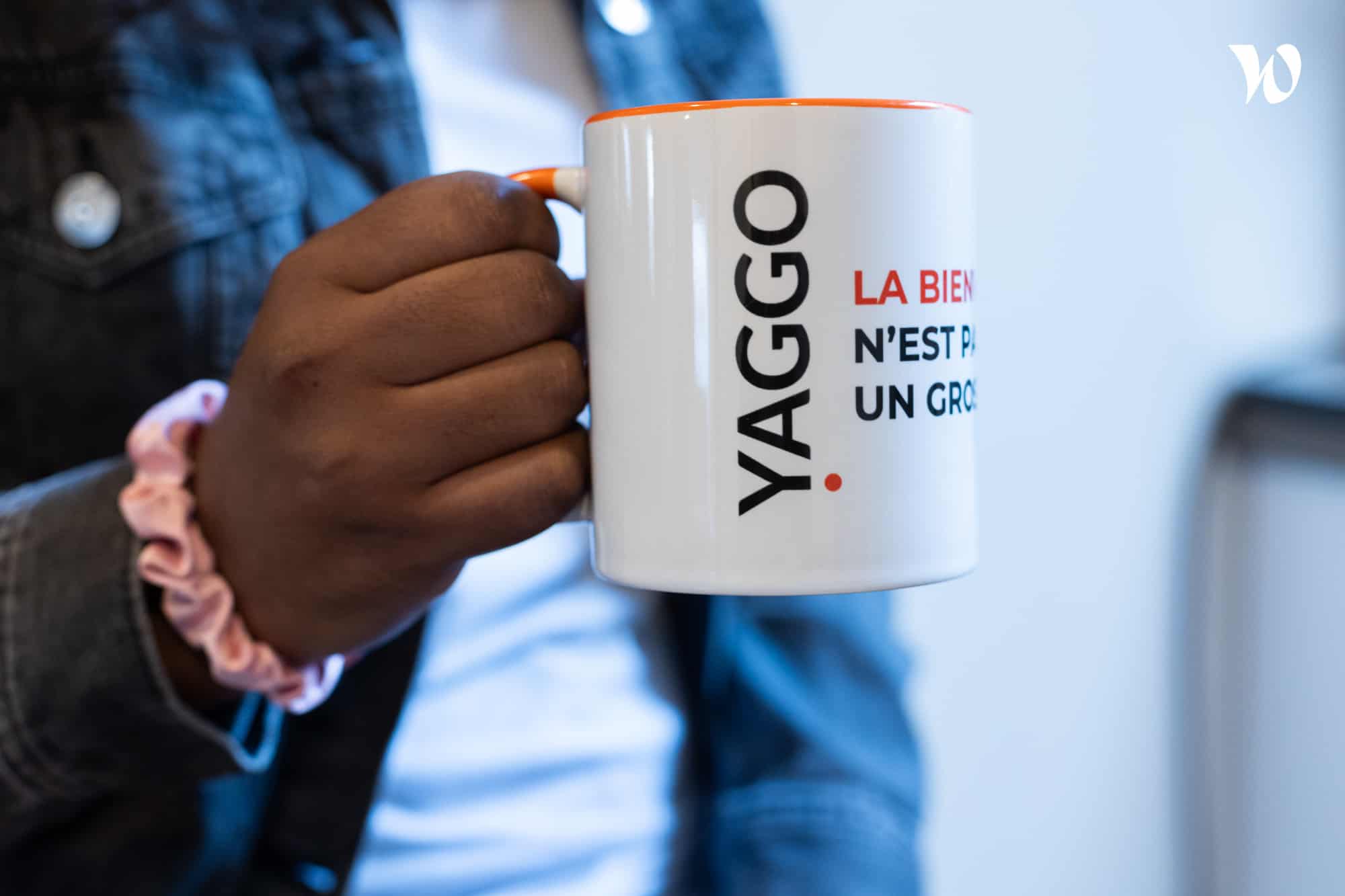 Main tenant une tasse blanche avec le logo YAGGO et le texte partiel « LA BIEN... N'EST PA... UN GRO... » en noir et rouge.