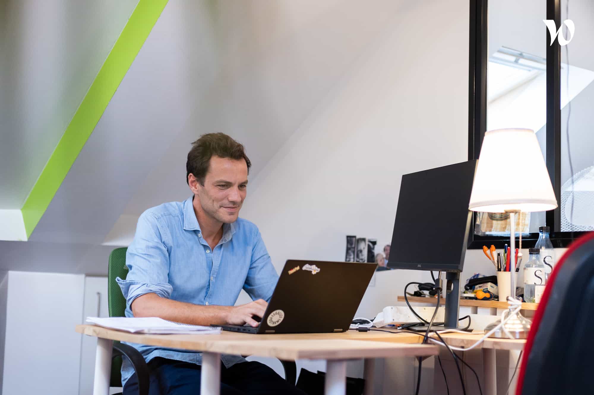 Un homme souriant en chemise bleue assis à un bureau, travaillant sur un ordinateur portable noir dans un bureau lumineux.