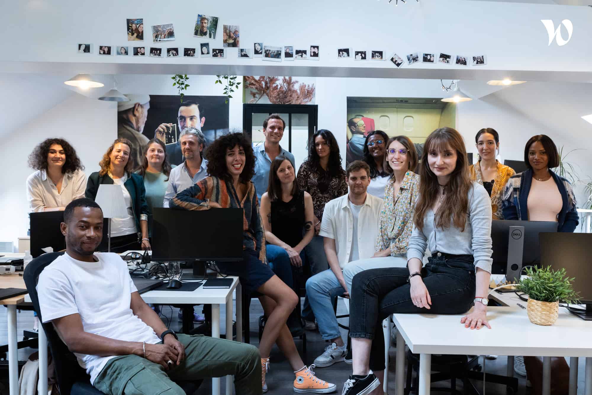 Groupe de dix-sept personnes souriantes posant dans un bureau moderne avec des ordinateurs et des photos accrochées au mur.