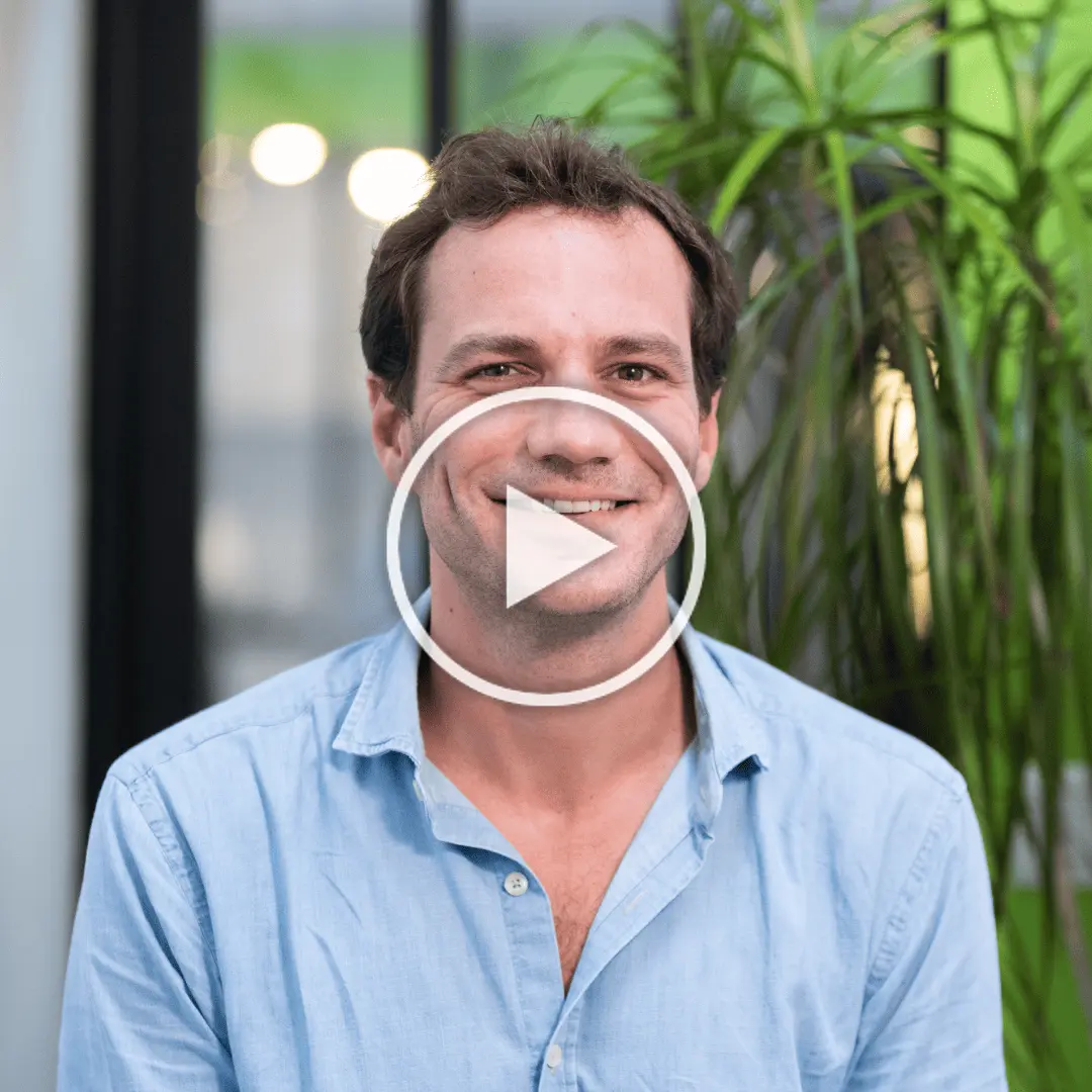 Homme souriant portant une chemise bleu clair avec des plantes en arrière-plan flou.