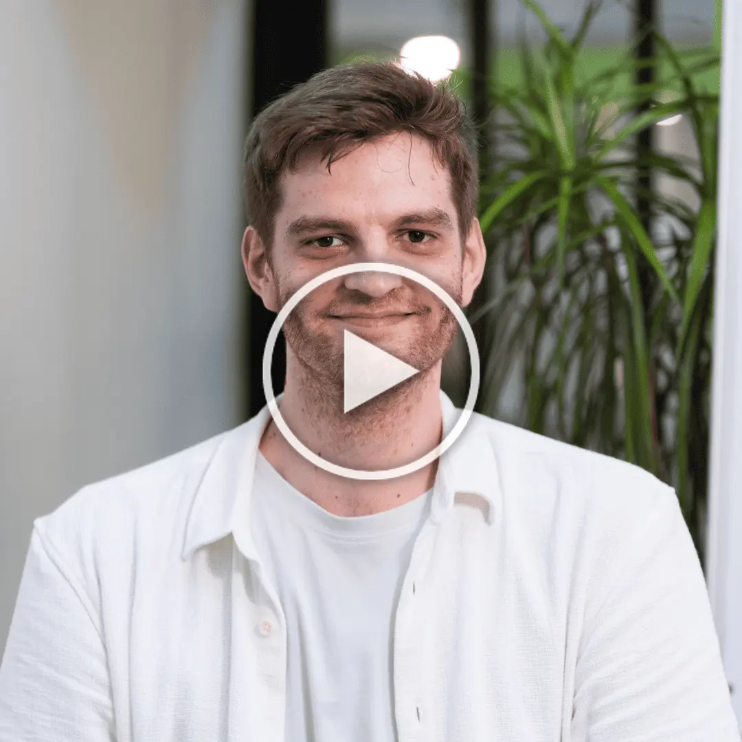 Un homme souriant avec une barbe légère porte une chemise blanche, avec un décor intérieur et une plante en arrière-plan.