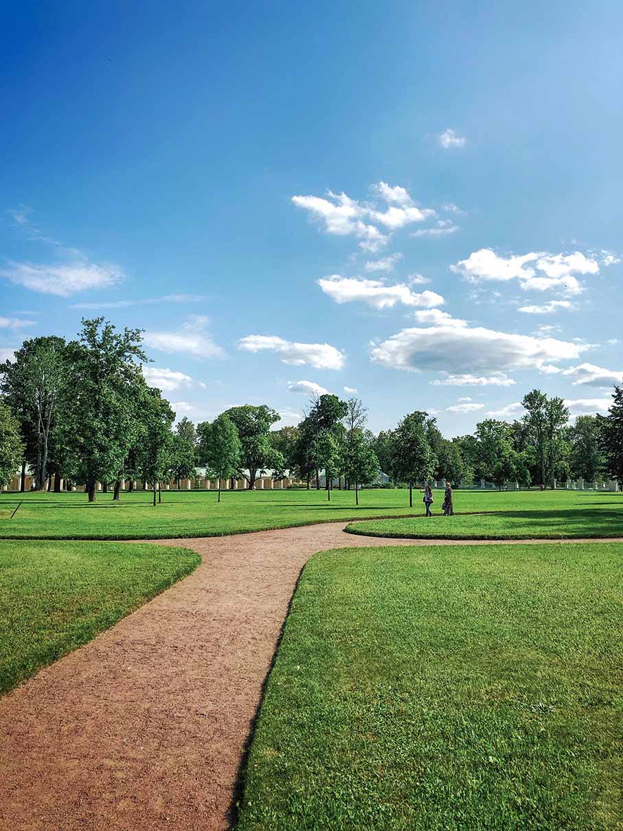 Park mit einem geschwungenen Kiesweg, umgeben von gepflegtem Gras und Bäumen unter blauem Himmel mit einigen Wolken.