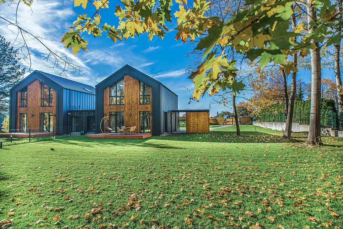Zwei moderne Häuser mit Holz- und Metallfassaden stehen auf einem großen, grünen Rasen mit herabgefallenen Herbstblättern, umgeben von Bäumen unter einem blauen Himmel.